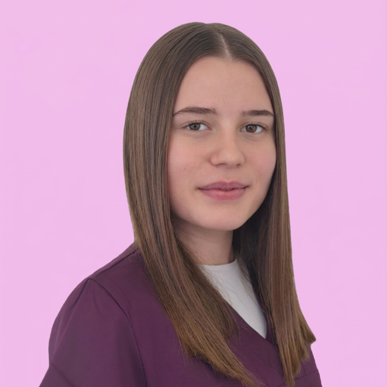 Frau mit langen, dunklen Haaren lächelt, trägt scrubs in lila. Hintergrund beige.