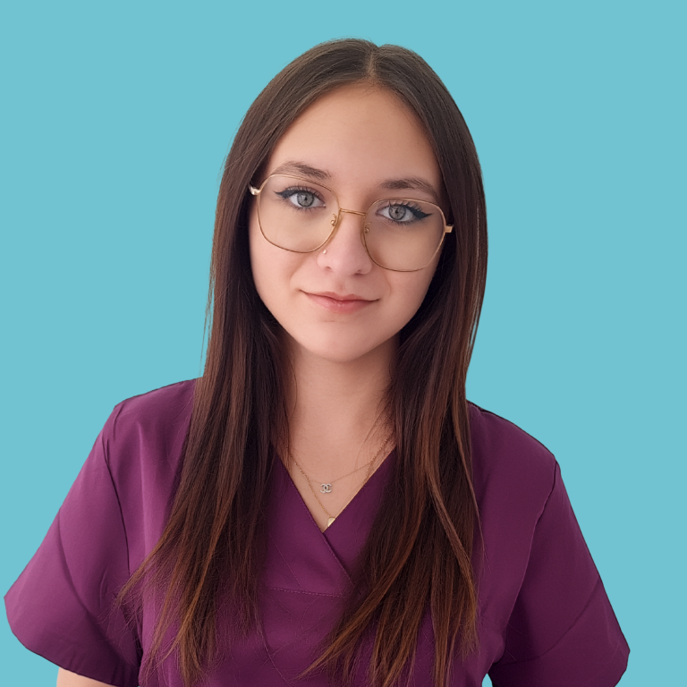 Frau mit langen, dunklen Haaren lächelt, trägt scrubs in lila. Hintergrund beige.