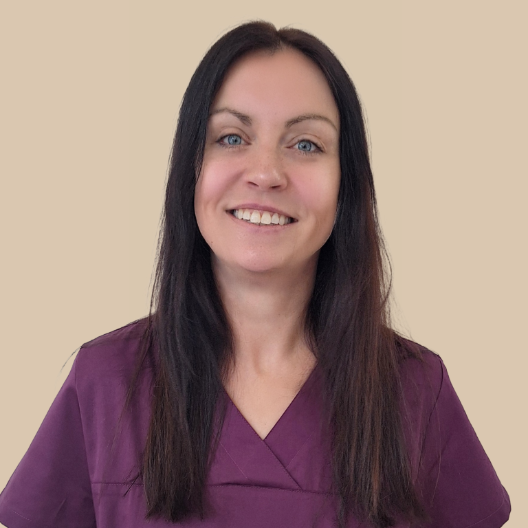 Frau mit langen, dunklen Haaren lächelt, trägt scrubs in lila. Hintergrund beige.