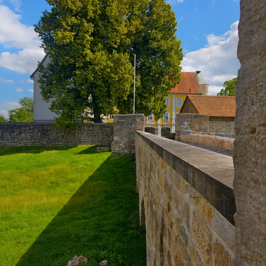 Mauer mit Sicht auf grüne Wiese und dem Schloss unter blauem Himmel mit Wolken.