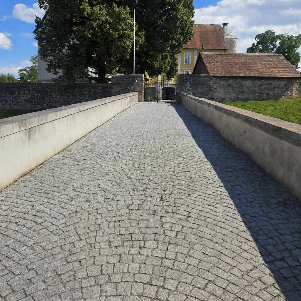 Pflasterweg zwischen Mauern, mit Wiesen und einem Schloss im Hintergrund.